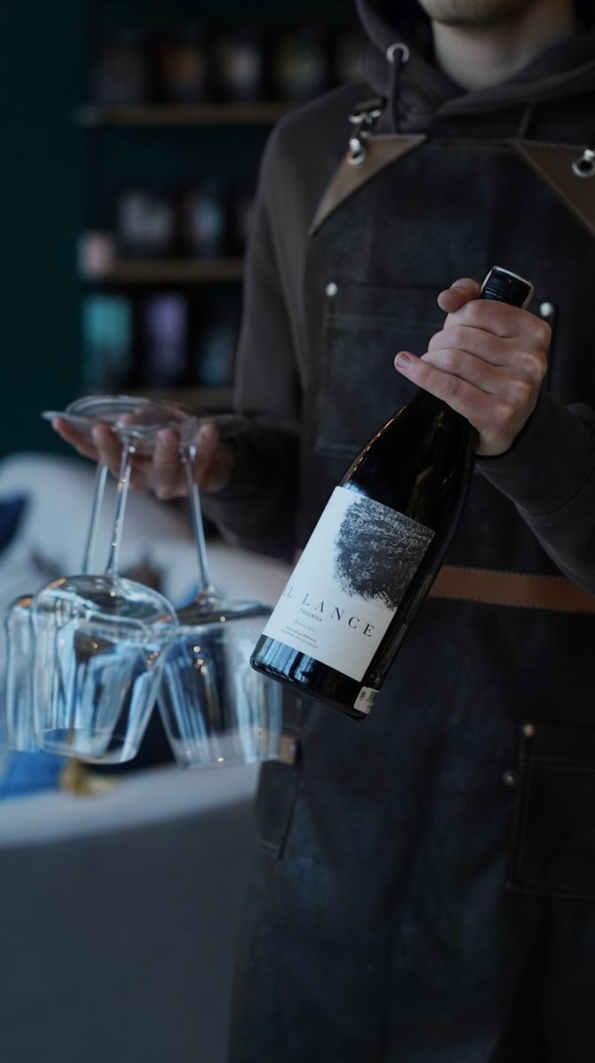 Photo by Anastasia Ilina-Makarova A person in an apron holds a wine bottle and glasses in an indoor setting.