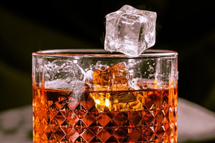 Photo by Nano Erdozain A detailed shot of whisky in a patterned glass with an ice cube mid-air, evoking elegance.
